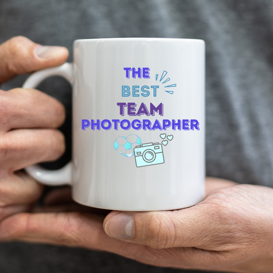 Text in blue saying the best team photographer with the image of a camera and heart with soccer ball print on a white mug.