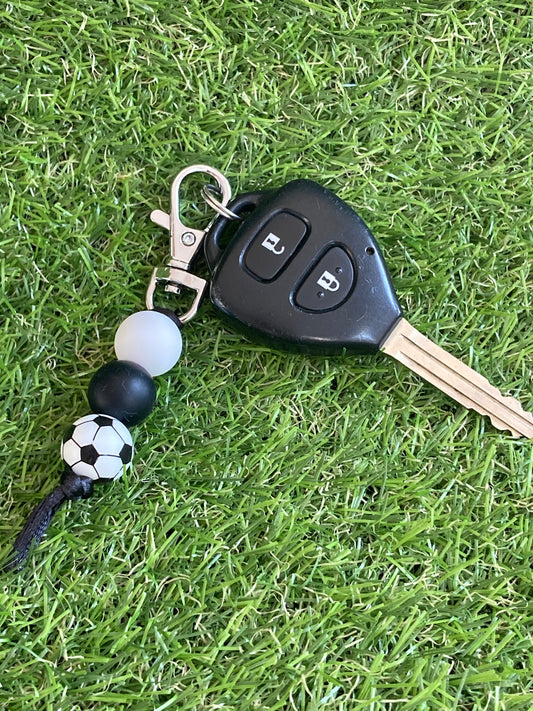 Beaded keychain that is black and white with a soccer ball bead attached to a key lying on grass.