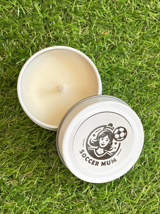 Candle in a white tin with a Soccer Mum logo on top laying on the grass.